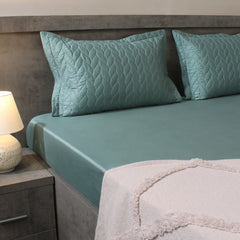 Teal bed sheets and pillows on a bed with a nightstand and lamp in the background.