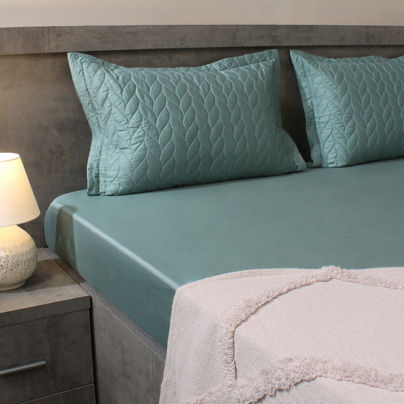 Teal bed sheets and pillows on a bed with a nightstand and lamp in the background.