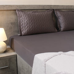 Gray quilted bedspread and pillows on a bed with a lamp on a nightstand.