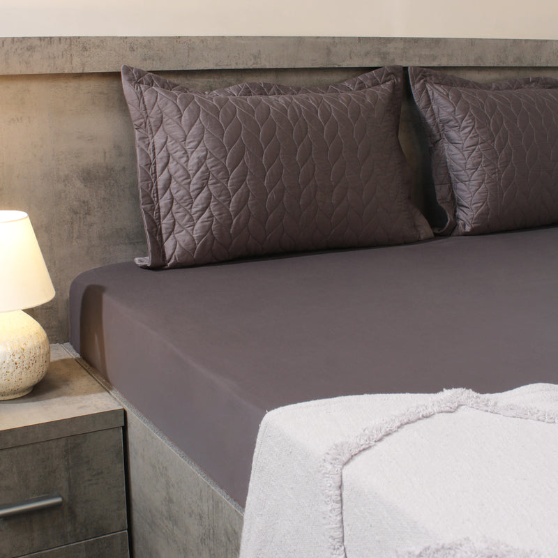Gray quilted bedspread and pillows on a bed with a lamp on a nightstand.